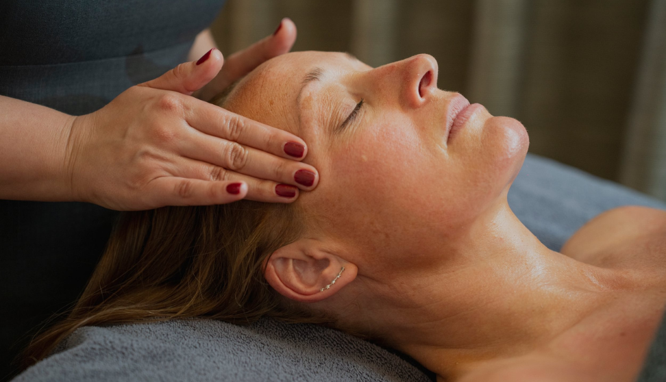 A woman is lying down with her eyes closed, receiving a relaxing massage on her forehead and temples from a therapist whose hands are visible. The setting appears calm and serene, suggesting a spa or wellness environment.