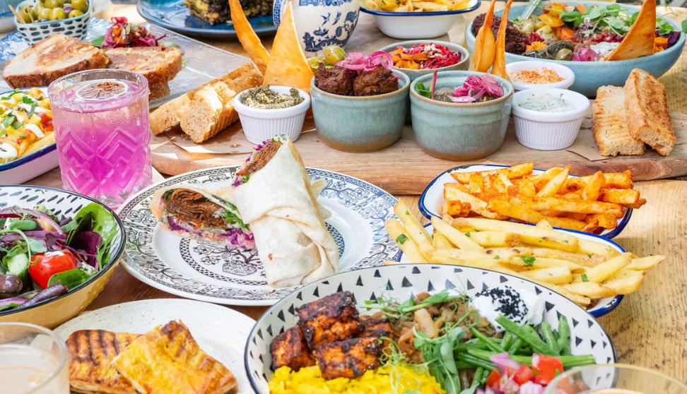 A colourful table filled with a wide variety of vegetarian and vegan-style dishes, including wraps, grilled tofu or paneer, salads, dips, fries and bread, all served in patterned bowls and plates on a wooden surface, with bright pink and light drinks in patterned glasses, creating a vibrant, abundant sharing feast.