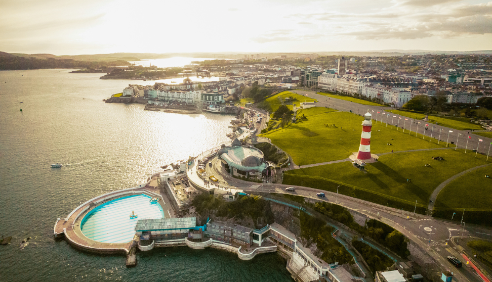 Aerial view of Plymouth Hoe at sunset, showing the outdoor lido, curved seafront road, wide green park, and red‑and‑white lighthouse beside the coastline.