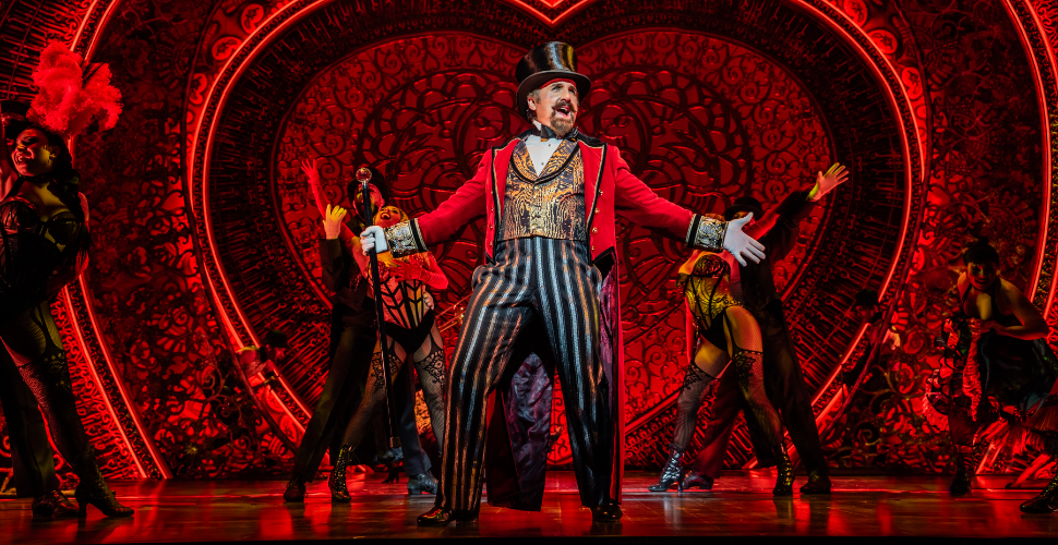 Ringmaster leading a high-energy Moulin Rouge–style musical number on stage, surrounded by dancers in red and black costumes against an ornate heart-shaped backdrop.