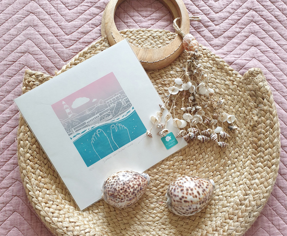 A seaside lino art print sits with some shells on a circular wicker bag