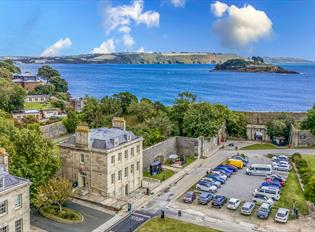 Aerial view of a coastal town with historic stone buildings and lush trees. A parking lot sits near the blue ocean, with an island in the distance.