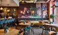 Stylish café with a checkered floor and vibrant wall art depicting abstract figures. Cosy seating includes teal bar stools and a mustard sofa.