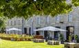 Historic stone building with a grassy courtyard, shaded outdoor seating, and large umbrellas. Trees provide shade, creating a serene, inviting atmosph