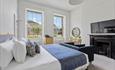 Bright, airy bedroom with a large bed adorned with white and blue linens, a dark sofa, a fireplace, two large windows, and a desk. Calm and inviting.