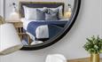 Reflection of a cosy bedroom in a round mirror. The bed has white and blue pillows, wood headboard, and wall lamps. Nearby, a desk holds a lamp, hat,